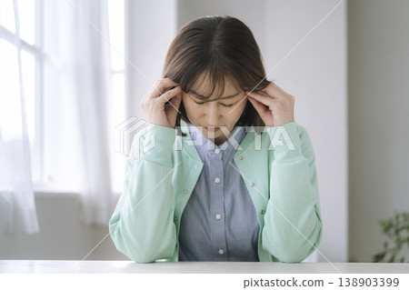A woman in her late 40s or early 50s rests both elbows on the table and holds her head in her hands. A woman in her late 40s or early 50s rests both elbows on the table and holds her head in her hands. 138903399