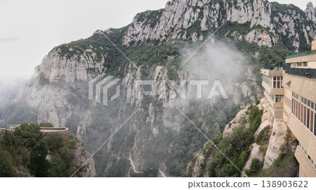 A scenic view of the rugged Montserrat mountains with mist swirling around the peaks and a building perched on the cliffside A scenic view of the rugged Montserrat mountains with mist swirling around the peaks and a building perched on the cliffside 138903622