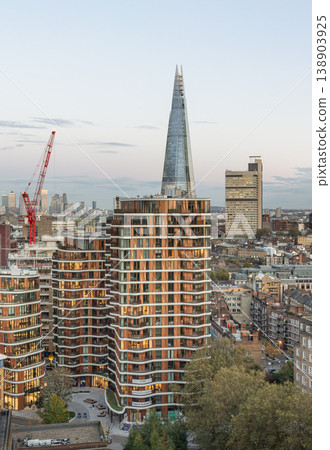 The shimmering glass spire of The Shard pierces the soft evening sky, standing as a majestic centerpiece amidst the urban sprawl of London. In the foreground, the undulating, golden-lit curves of the The shimmering glass spire of The Shard pierces the soft evening sky, standing as a majestic centerpiece amidst the urban sprawl of London. In the foreground, the undulating, golden-lit curves of the 138903925