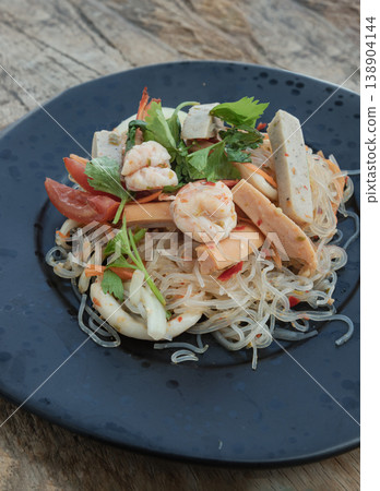 Presented on a matte black plate, this vibrant Yum Woon Sen features shimmering glass noodles tossed with succulent shrimp, tender squid, and savory pork sausage. Aromatic celery and bright tomatoes Presented on a matte black plate, this vibrant Yum Woon Sen features shimmering glass noodles tossed with succulent shrimp, tender squid, and savory pork sausage. Aromatic celery and bright tomatoes 138904144