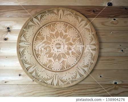 A circular wooden platter with hand carved floral and geometric patterns sits on a wooden wall. This fine craftsmanship shows traditional techniques used in crafting decor items. 138904276