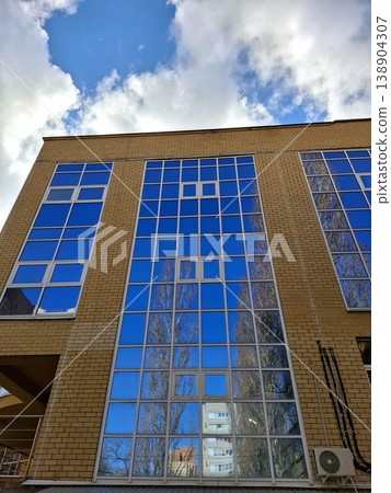 A low angle view shows a modern brick building with large blue glass windows. The windows reflect nearby trees and the sky with clouds. The scene takes place in an urban setting under clear daylight. A low angle view shows a modern brick building with large blue glass windows. The windows reflect nearby trees and the sky with clouds. The scene takes place in an urban setting under clear daylight. 138904307