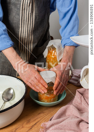 A 53-year-old man prepares to ferment cabbage and carrots in his kitchen. He carefully places a jar filled with vegetables on a bowl. The workspace shows various tools 138906891