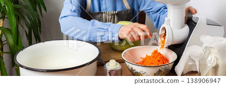 A 53-year-old man is in his kitchen preparing cabbage and carrots for fermentation. He uses a machine to shred the vegetables before placing them in a bowl, banner 138906947