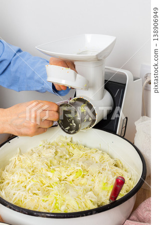 A 53-year-old man prepares cabbage and carrots for fermentation in his kitchen. He uses a machine to shred the vegetables while surrounded by kitchen tools and ingredients 138906949
