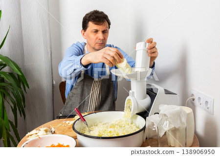 A 53-year-old man prepares cabbage and carrots for fermentation in his kitchen. He uses a machine to shred the vegetables while surrounded by kitchen tools and ingredients 138906950