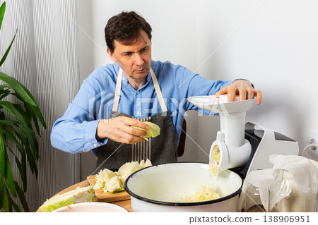 A 53-year-old man prepares cabbage and carrots for fermentation in his kitchen. He uses a machine to shred the vegetables while surrounded by kitchen tools and ingredients 138906951