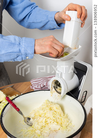 A 53-year-old man prepares cabbage and carrots for fermentation in his kitchen. He uses a machine to shred the vegetables while surrounded by kitchen tools and ingredients 138906952
