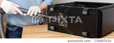 A 53-year-old man uses a vacuum to clean dust from a computer system unit on a wooden table in a home office. The setting is tidy and organized, banner 138907016