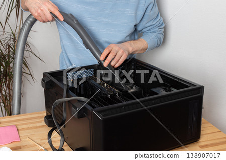 A 53-year-old man uses a vacuum attachment to remove dust from a computer system unit in a home office. The room is well lit and organized for the task 138907017