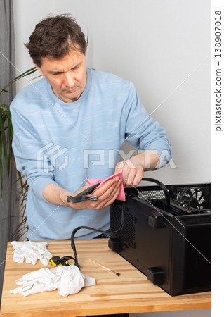 A 53-year-old man uses a cloth to clean dust from a computer system unit. He works at a wooden table in a room with plants nearby. Tools are set out for the task A 53-year-old man uses a cloth to clean dust from a computer system unit. He works at a wooden table in a room with plants nearby. Tools are set out for the task 138907018