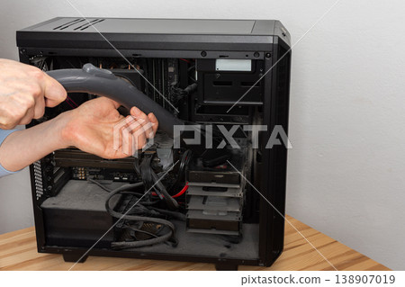 A 53-year-old man uses a vacuum attachment to remove dust from a computer system unit in a home office. The room is well lit and organized for the task 138907019