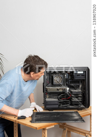A 53-year-old man removes dust from a computer system unit on a wooden table. He is focused on cleaning the inside of the computer in a well-lit room 138907020