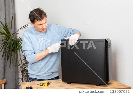 A 53-year-old man wears white gloves while cleaning dust from a computer system unit on a wooden table in a home workspace during the day 138907021