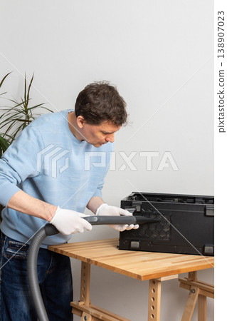 A 53-year-old man uses a vacuum to clean dust from a computer system unit on a wooden table in a home office. The setting is tidy and organized A 53-year-old man uses a vacuum to clean dust from a computer system unit on a wooden table in a home office. The setting is tidy and organized 138907023