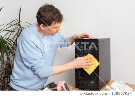 A 53-year-old man uses a cloth to clean dust from a computer system unit. He works at a wooden table in a room with plants nearby. Tools are set out for the task A 53-year-old man uses a cloth to clean dust from a computer system unit. He works at a wooden table in a room with plants nearby. Tools are set out for the task 138907024