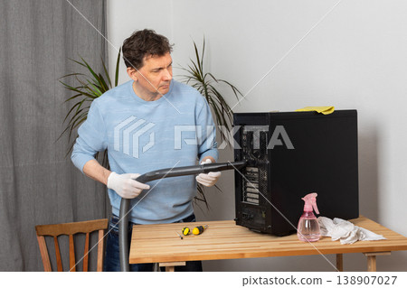 A 53-year-old man cleans dust from a computer system unit with a vacuum cleaner in a tidy room. He carefully removes dirt to keep the device running well A 53-year-old man cleans dust from a computer system unit with a vacuum cleaner in a tidy room. He carefully removes dirt to keep the device running well 138907027