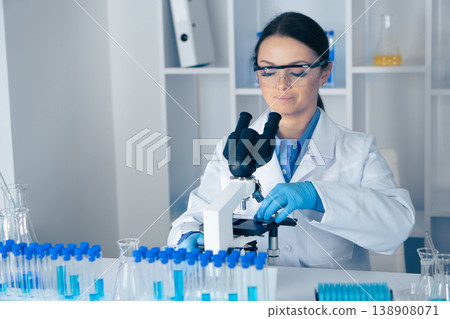 A dedicated female scientist examines samples through a microscope, surrounded by test tubes and advanced lab equipment, utilizing the latest technology for research and discovery A dedicated female scientist examines samples through a microscope, surrounded by test tubes and advanced lab equipment, utilizing the latest technology for research and discovery 138908071