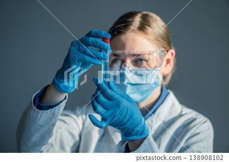 A female scientist wearing gloves, mask, and goggles carefully studies a test tube filled with blue solution in a modern lab. Laboratory research highlights medical and scientific progress A female scientist wearing gloves, mask, and goggles carefully studies a test tube filled with blue solution in a modern lab. Laboratory research highlights medical and scientific progress 138908102