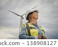 A highly skilled female engineer, dressed in safety gear, stands confidently before a towering wind turbine, which symbolizes both progress and innovation in the vital field of renewable energy 138908127
