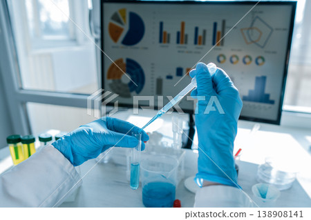 Closeup of laboratory research as a gloved hand uses a pipette to move blue liquid into a test tube. Digital graphs and scientific charts are visible on a screen, highlighting data analysis Closeup of laboratory research as a gloved hand uses a pipette to move blue liquid into a test tube. Digital graphs and scientific charts are visible on a screen, highlighting data analysis 138908141