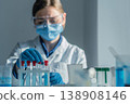 A focused female researcher handles test tubes with blue solution, wearing safety gear in a contemporary laboratory. The scene highlights innovation, science, and healthcare advancements 138908146