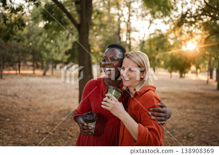 Two happy multi-ethnic friends laughing and hugging in a park at sunset, drinking coffee Two happy multi-ethnic friends laughing and hugging in a park at sunset, drinking coffee 138908369
