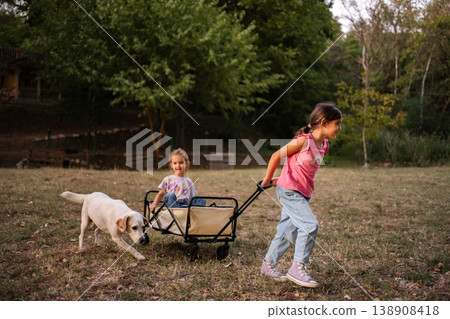 Girl pulling her twin sister in a cart with their dog running alongside 138908418