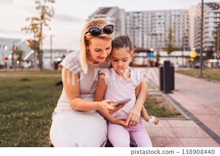 Mother and daughter looking at smartphone sitting on bench in urban park Mother and daughter looking at smartphone sitting on bench in urban park 138908440