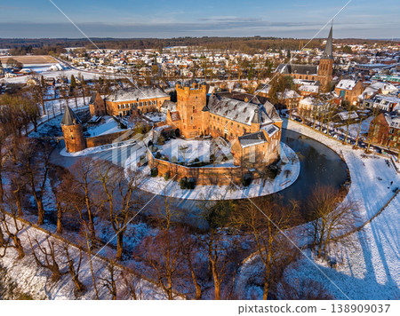 Aerial view of medieval brick castle in snow-covered town with large moat, church and forest in winter under sunny blue sky. Castle in winter. Aerial view of medieval brick castle in snow-covered town with large moat, church and forest in winter under sunny blue sky. Castle in winter. 138909037