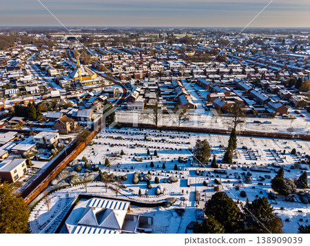 Aerial view of a snowy cemetery and residential district in a European town. Winter cityscape with snow-covered rooftops, church, and rows of graves under a clear sunny sky. Aerial view of a snowy cemetery and residential district in a European town. Winter cityscape with snow-covered rooftops, church, and rows of graves under a clear sunny sky. 138909039