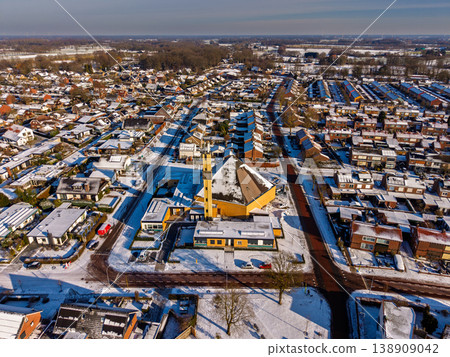 High-angle aerial view of a modern church and residential neighborhood covered in snow. A sunny winter morning in a European town with white rooftops, long shadows, and clear blue skies. High-angle aerial view of a modern church and residential neighborhood covered in snow. A sunny winter morning in a European town with white rooftops, long shadows, and clear blue skies. 138909042