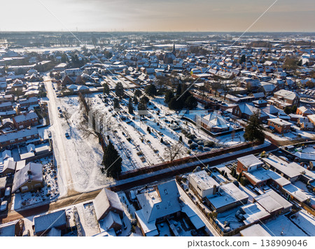 Aerial winter panorama of a snow-covered town in Europe featuring a central cemetery, a church with a spire, and residential neighborhoods under a bright, hazy sun with long blue shadows. Aerial winter panorama of a snow-covered town in Europe featuring a central cemetery, a church with a spire, and residential neighborhoods under a bright, hazy sun with long blue shadows. 138909046