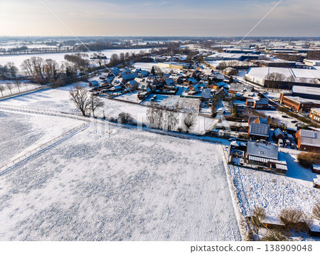 High-angle aerial view of a snowy European village and industrial zone. Residential houses and fields covered in fresh white snow under a bright winter sun. Scenic cold weather landscape. High-angle aerial view of a snowy European village and industrial zone. Residential houses and fields covered in fresh white snow under a bright winter sun. Scenic cold weather landscape. 138909048