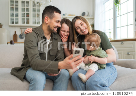 Happy family talk on video call with friends family. Mother father two children holding smartphone talking speaking online. Family mom dad parents baby girls kids smiling laughing enjoying moment 138910274
