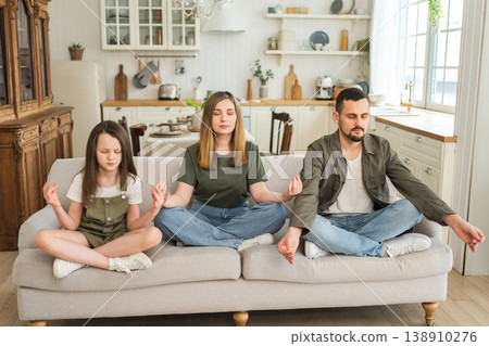 Yoga mindfulness meditation emotional wellbeing. Happy family practicing yoga at home Mother father daughter sitting in lotus pose meditating relaxing on couch Family doing breathing practice together 138910276