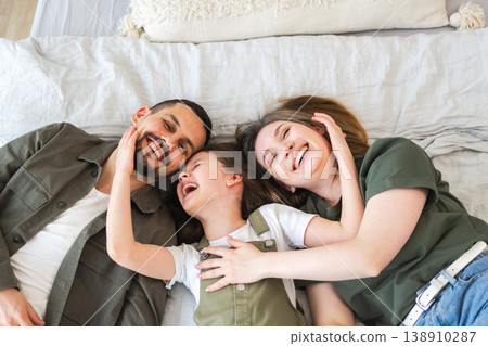 Happy family at home. Mother father children daughter relaxing in bed at home indoor. Mom dad parents kid girl relax playing hugging having fun together. Family smiling laughing enjoying tender moment 138910287