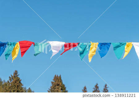 Colorful flags hang in a clear blue sky over trees in a sunny environment during the day Colorful flags hang in a clear blue sky over trees in a sunny environment during the day 138911221