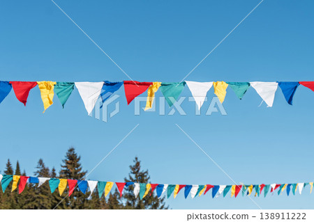 Colorful flags hang against blue sky with trees in background during outdoor celebration scene 138911222