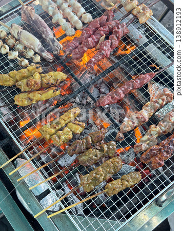 Assortment of skewered meats and seafood grilling over hot coals at an outdoor stall Assortment of skewered meats and seafood grilling over hot coals at an outdoor stall 138912193