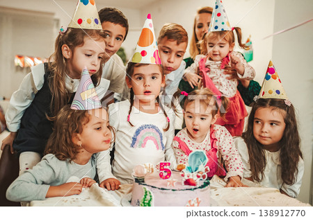 Group of children gather to celebrate a birthday with hats and a cake while preparing to blow out candles Group of children gather to celebrate a birthday with hats and a cake while preparing to blow out candles 138912770