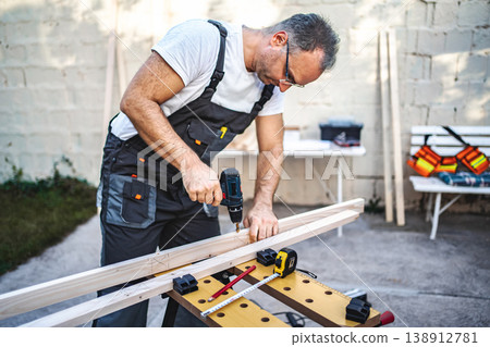 Mature male carpenter using electric drill on a wooden plank outdoors 138912781