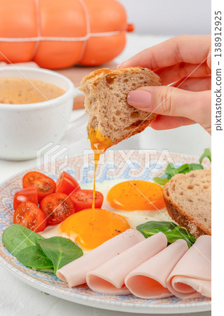 Breakfast plate with fried eggs, bread and turkey slices, hand dipping bread into runny yolk 138912925