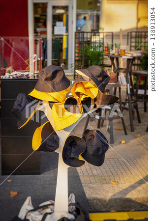 Display of bucket hats in black and yellow on stand outside street shop with clothing store background and outdoor retail setting. Fashion retail, street market, summer accessory, urban shopping Display of bucket hats in black and yellow on stand outside street shop with clothing store background and outdoor retail setting. Fashion retail, street market, summer accessory, urban shopping 138913054