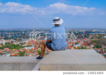 Young man sitting on concrete ledge overlooking city skyline with buildings, roofs and streets under sky, travel scene with urban perspective. Concept of travel, tourism lifestyle, reflections on life 138913064
