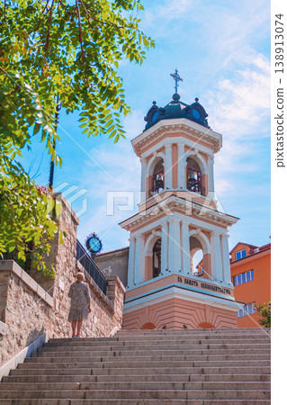 Plovdiv bell tower rising above stone stairs with person walking upward framed by green tree leaves and blue sky in historic city center. Travel destination, heritage tourism, city landmark Plovdiv bell tower rising above stone stairs with person walking upward framed by green tree leaves and blue sky in historic city center. Travel destination, heritage tourism, city landmark 138913074