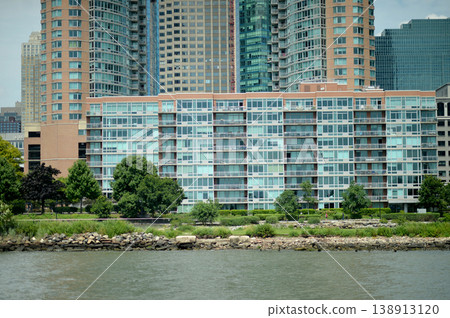 Riverfront Apartment Scene. Contemporary Dwelling Featuring Green Lawns And Cityscape Backdrop Riverfront Apartment Scene. Contemporary Dwelling Featuring Green Lawns And Cityscape Backdrop 138913120