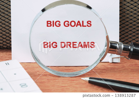 A magnifying glass focuses on the text Big Goals Big Dreams written on a white paper, placed on a wooden desk with a pen and calculator 138913287