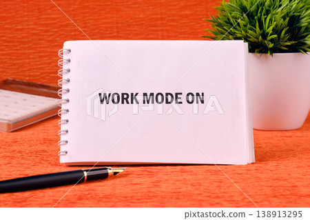 White spiral notebook with WORK MODE ON text on an orange surface. A black pen rests in front, a green potted plant sits to the side 138913295