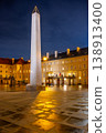 The Mrakotin Monolith stands tall at Prague Castle, surrounded by historical buildings, as warm light casts dramatic shadows on the cobblestone ground under the night sky. 138913400